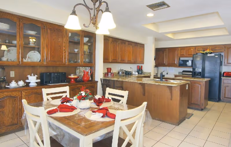 A kitchen and dining area with wooden cabinets and a tiled floor. The dining table is set with four white chairs, each place setting has a white plate with a red folded napkin. A floral centerpiece is on the table. The kitchen has granite countertops, a black refrigerator, a microwave, and various small appliances. A ceiling light fixture with three lamps hangs above the dining table.