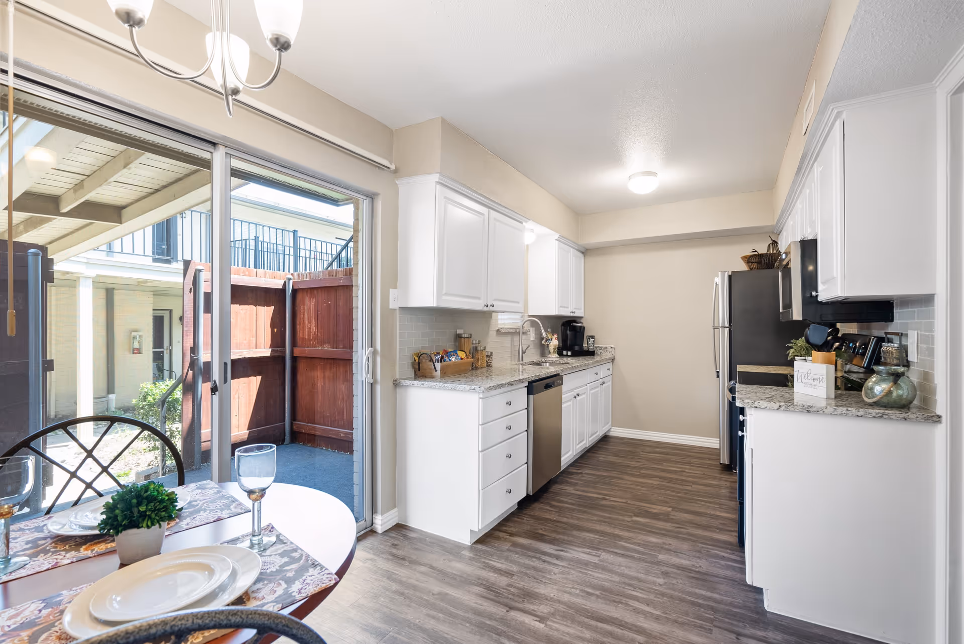 Bright kitchen with white cabinets, granite countertops, stainless steel appliances including a dishwasher, stove, microwave, and refrigerator. There is a small round dining table with two place settings and a potted plant near a sliding glass door that leads to a small outdoor patio area enclosed by a wooden fence.