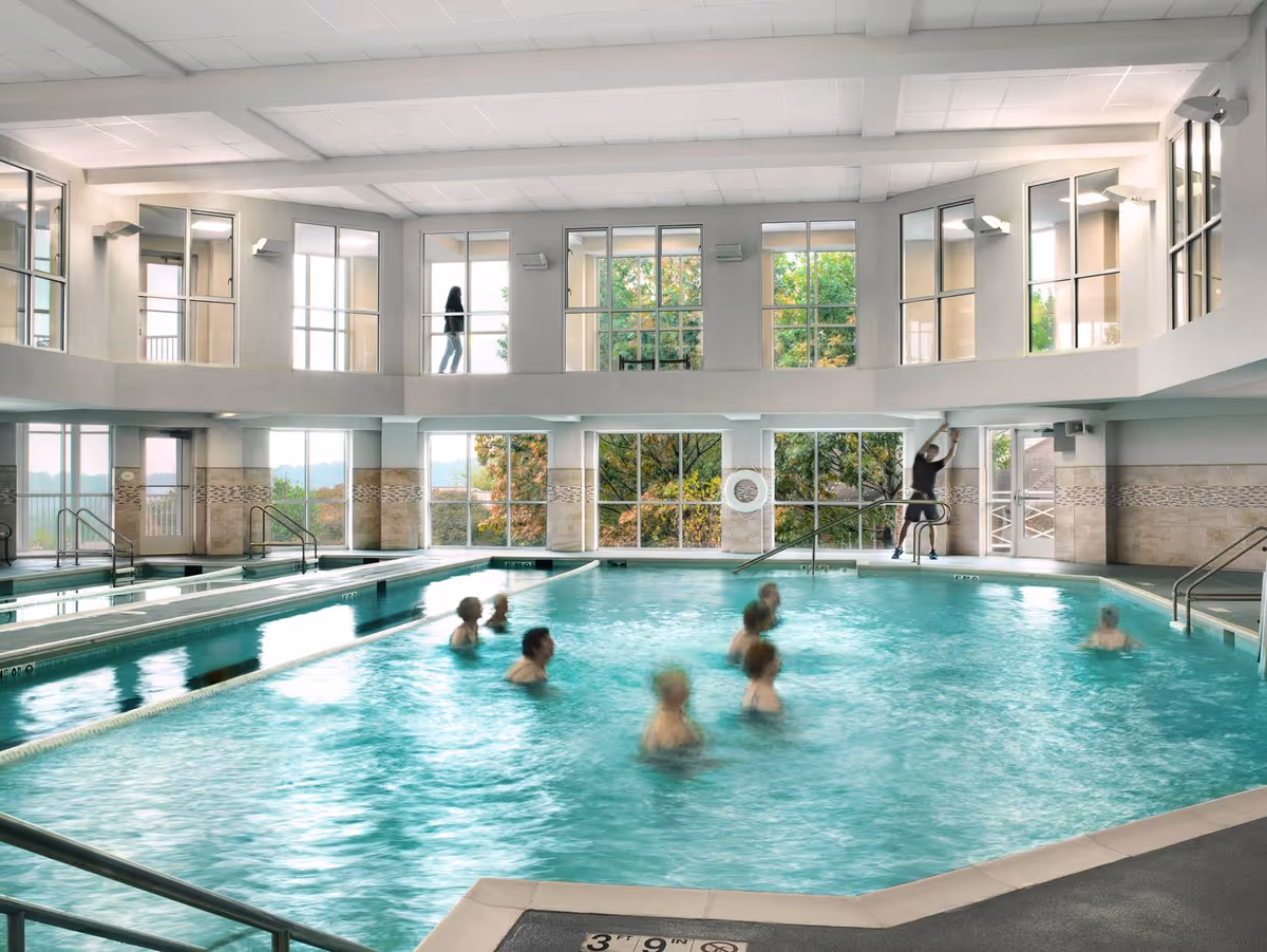 Indoor swimming pool with several people participating in a water exercise class. Large windows surround the pool area, allowing natural light to fill the space and providing views of trees outside. A person is seen walking on the upper level balcony overlooking the pool.