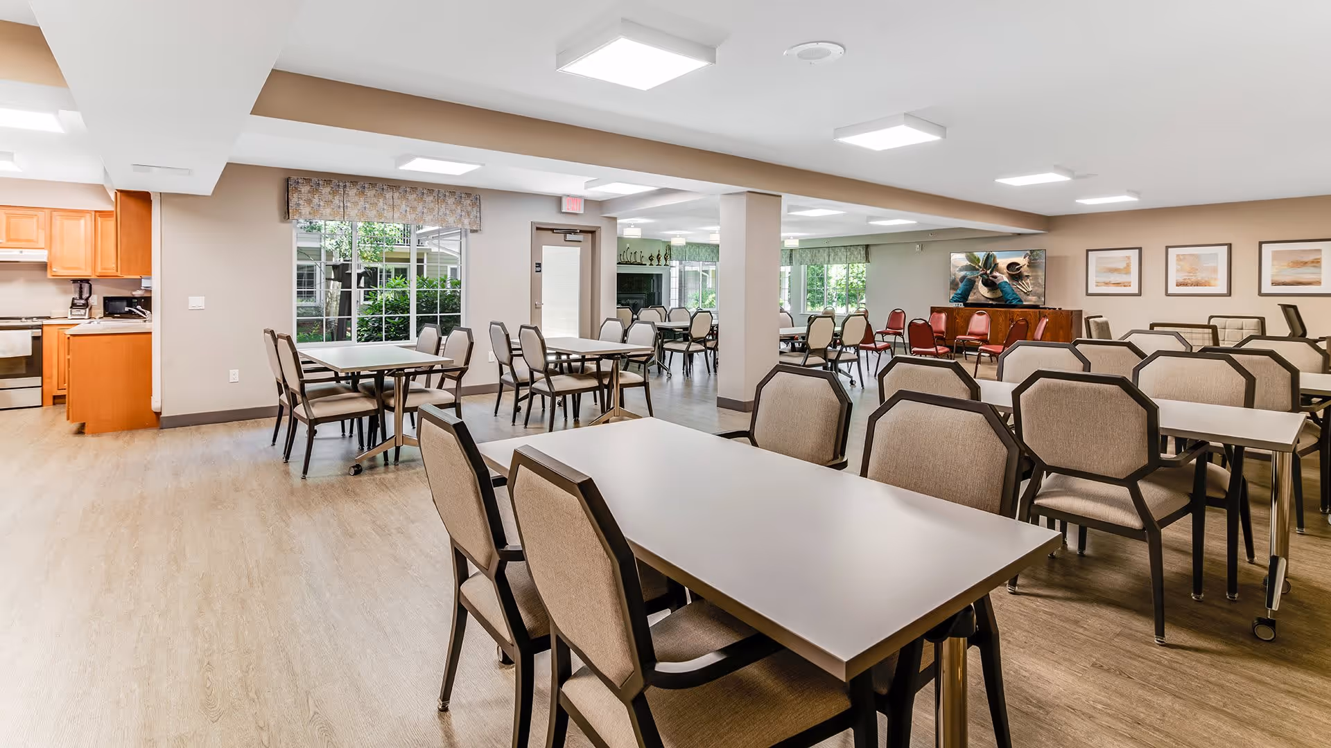 A spacious dining room with multiple tables and chairs arranged neatly. The room has light-colored walls, wood flooring, and large windows letting in natural light. There is a kitchen area visible on the left side with wooden cabinets and appliances. The far wall has framed artwork and a television mounted above a wooden cabinet. The room appears clean and well-lit, designed for communal dining.