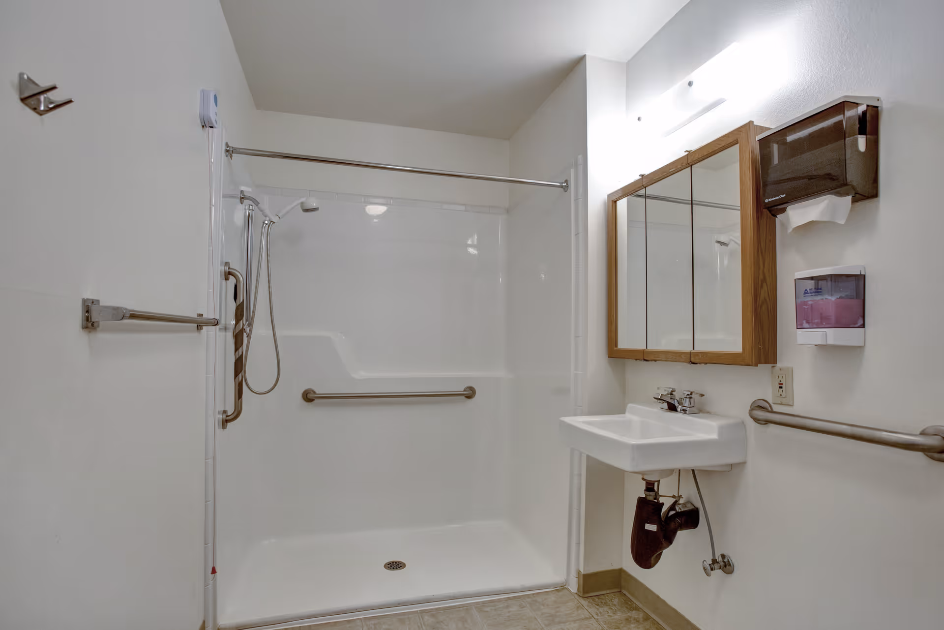 A clean, accessible bathroom with a walk-in shower featuring grab bars and a handheld showerhead. There is a wall-mounted sink with a mirror cabinet above it, a soap dispenser, and a paper towel dispenser on the wall. The walls are white and the floor has light-colored tiles.