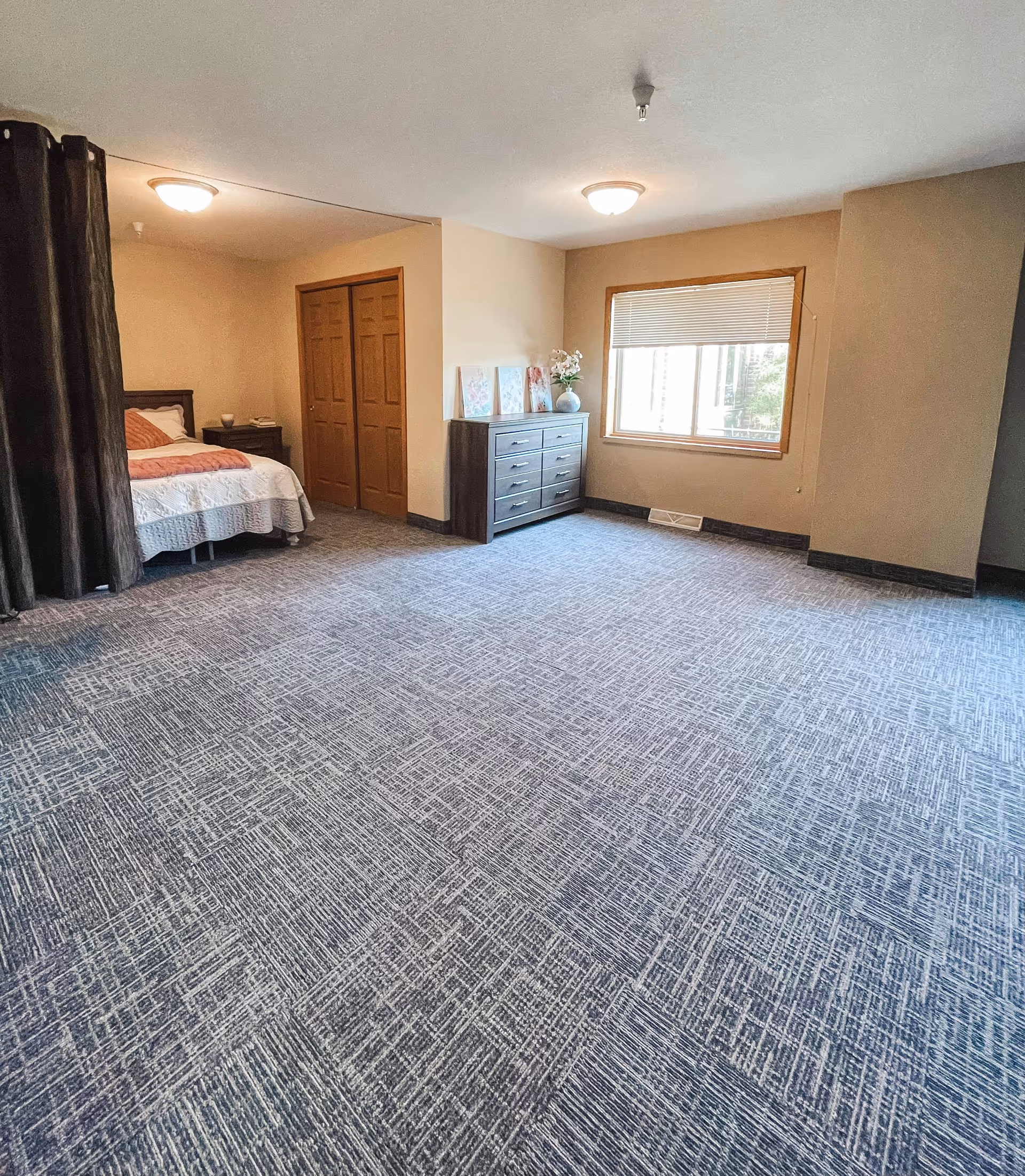 A spacious bedroom with a large window covered by a blind, a dark wooden dresser with decorative items on top, a bed partially visible behind a dark curtain, and a closet with double wooden doors. The room has beige walls and patterned carpet flooring.
