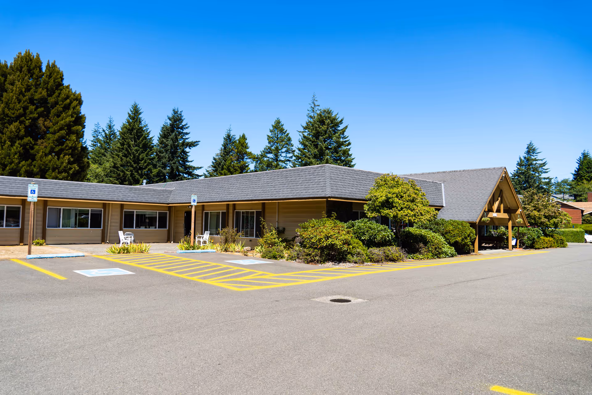 Exterior view of a single-story building with a gray roof and beige walls, surrounded by green bushes and trees under a clear blue sky. There are designated handicap parking spaces and yellow-striped no-parking zones in front of the building.
