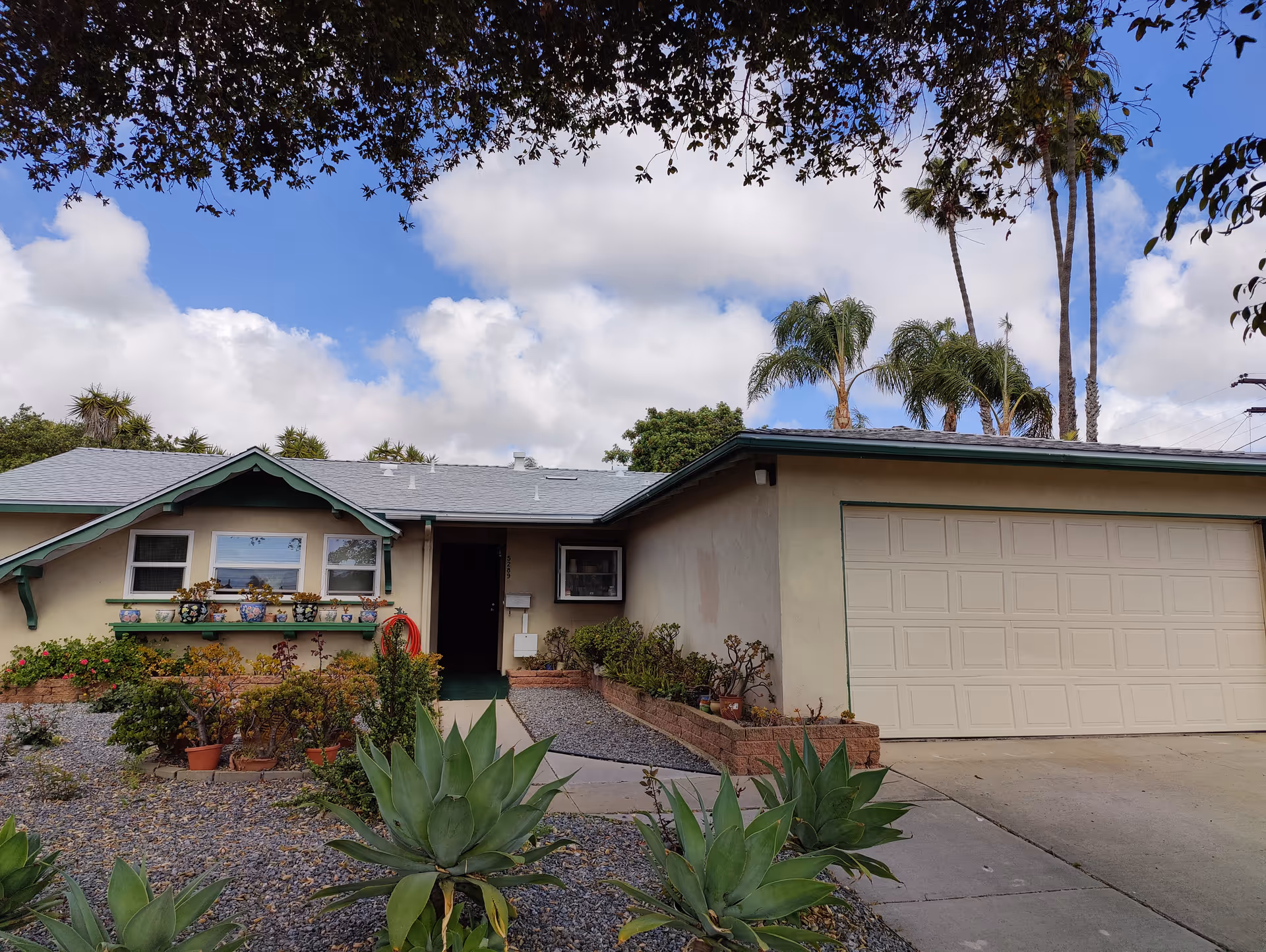 Front exterior view of a single-story house with a two-car garage, a small garden with various plants and succulents, and a pathway leading to the front door. The sky is partly cloudy with some palm trees visible in the background.
