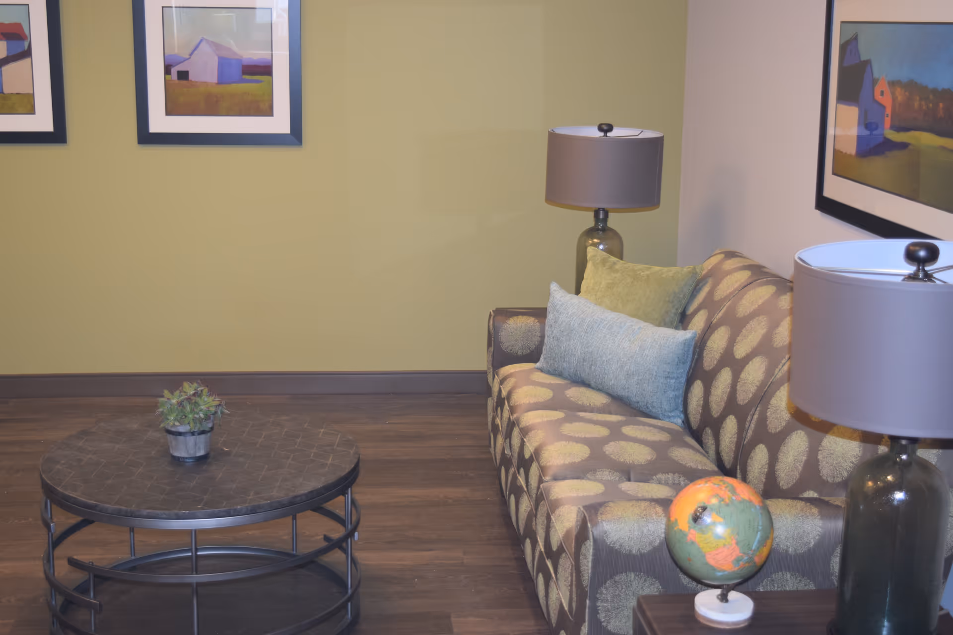A cozy living room area with a patterned sofa adorned with two cushions, a round coffee table with a small potted plant, two table lamps with gray shades, a small globe on a side table, and framed landscape paintings on the walls.