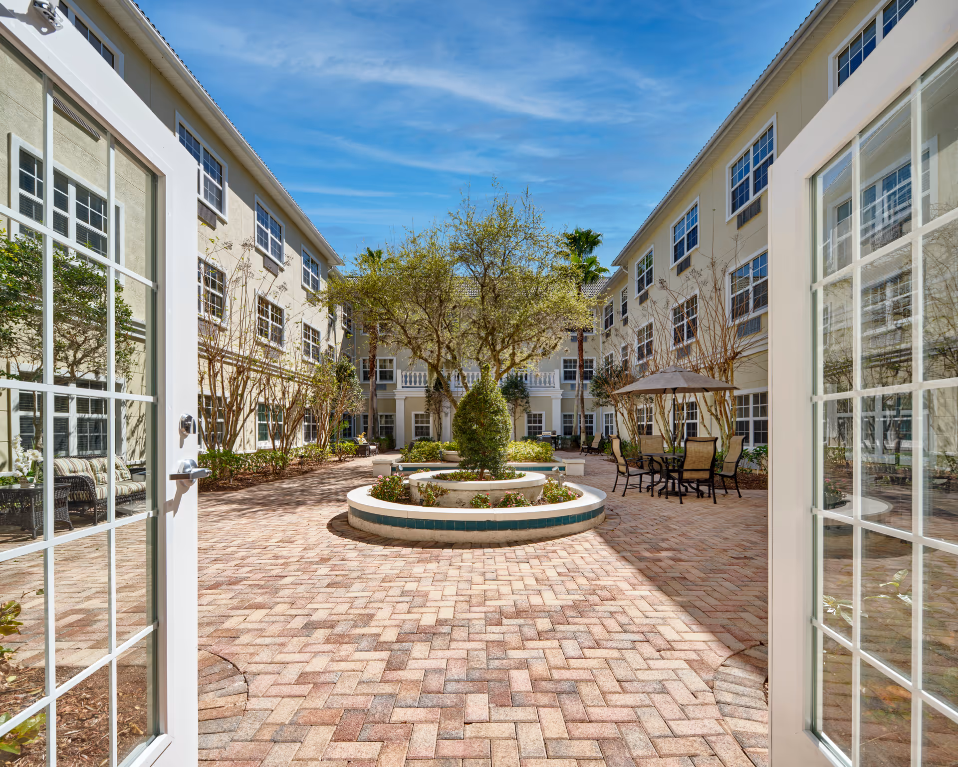 View through open double glass doors into a spacious outdoor courtyard at Colliers Assisted Living at Countryside. The courtyard features a circular raised planter with greenery and trees, surrounded by brick pavers. There are patio tables with umbrellas and chairs on the right side, and the courtyard is enclosed by a three-story building with many windows under a blue sky.