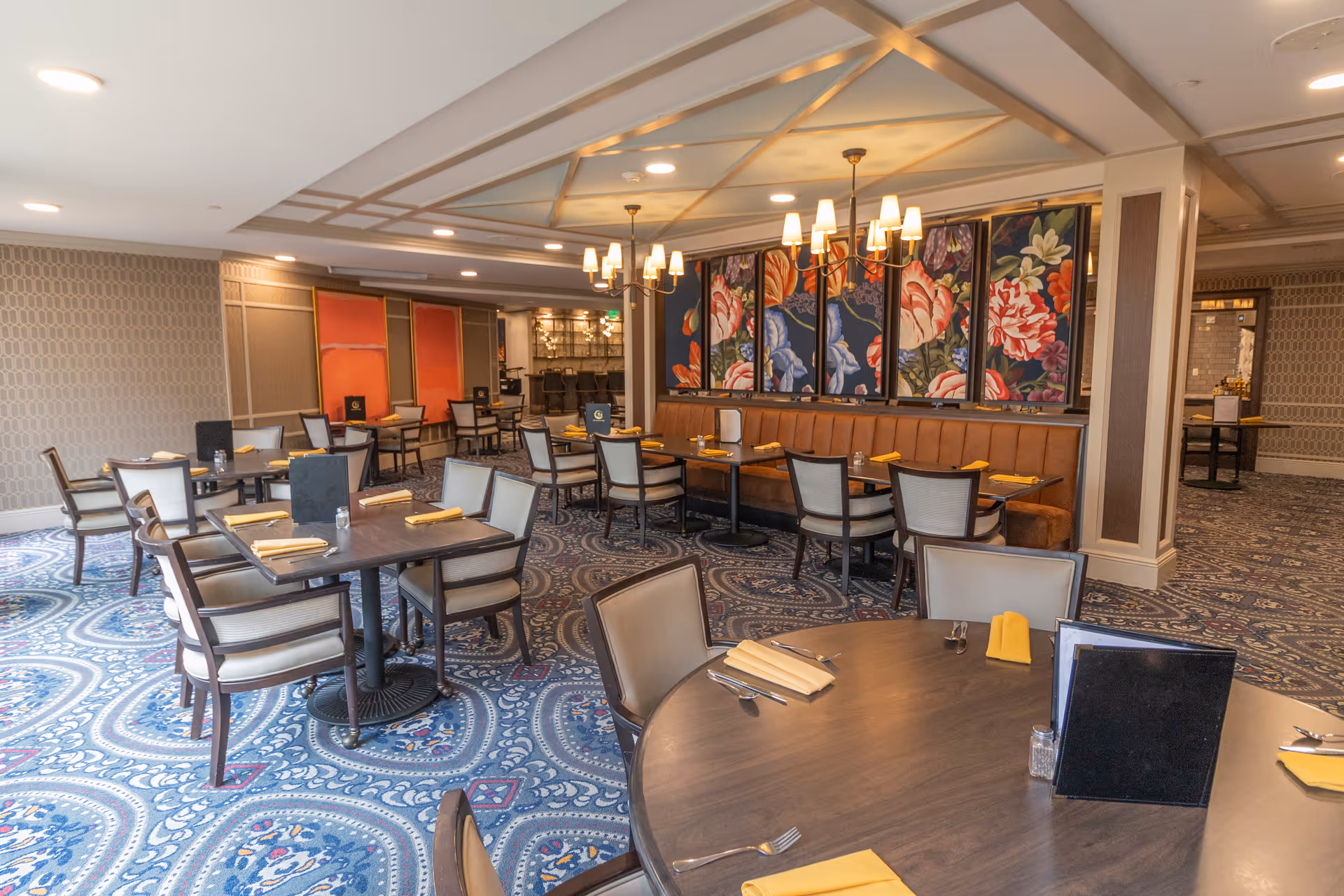 A spacious dining room with multiple tables and chairs arranged neatly. The tables are set with yellow napkins, silverware, and menus. The room features a patterned blue carpet, floral artwork on the walls, and warm lighting from ceiling fixtures.