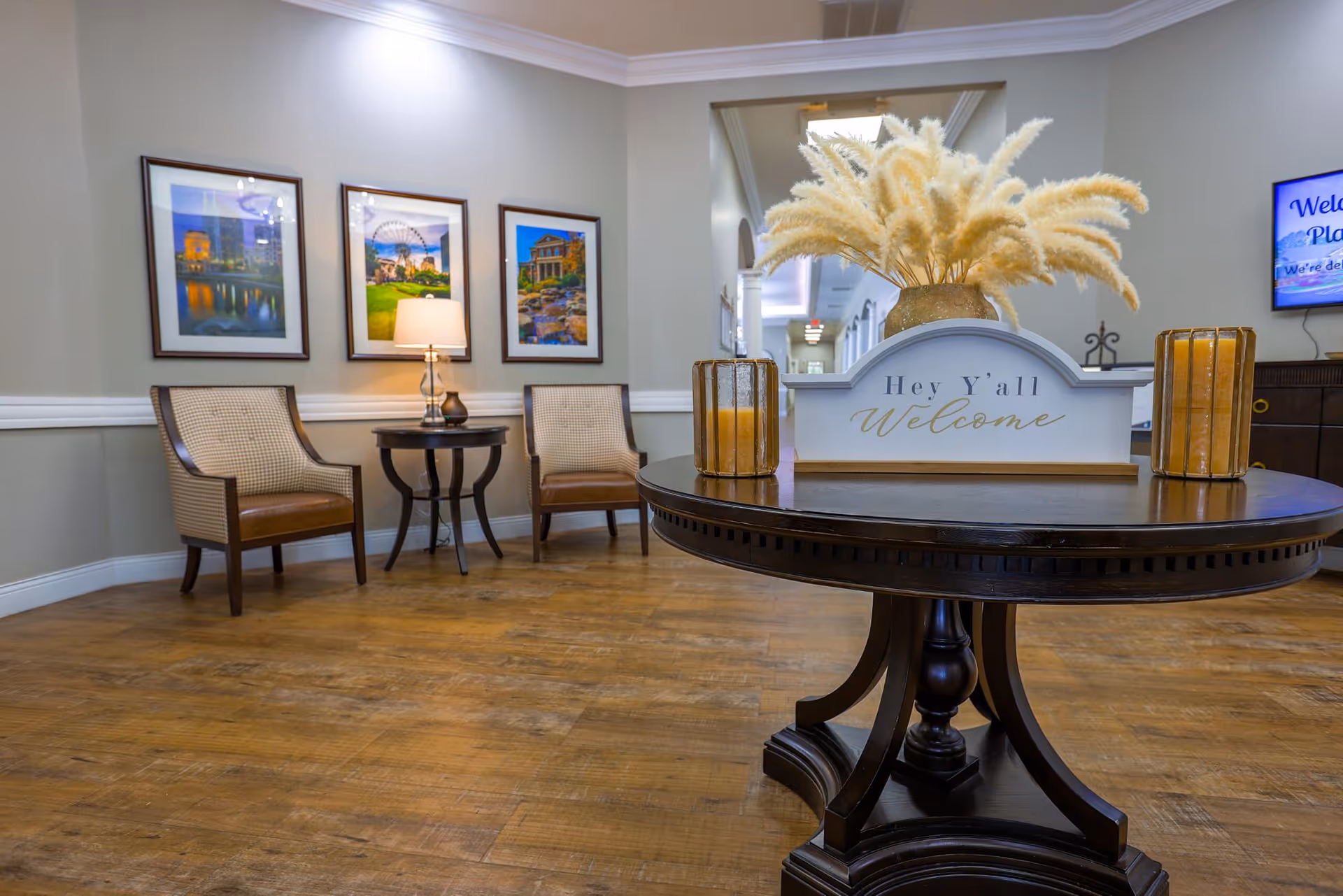 A welcoming seating area in a senior living facility with two cushioned chairs and a small round table with a lamp between them. A larger round wooden table in the foreground holds a decorative sign that says 'Hey Y'all Welcome' along with two candle holders and a vase with pampas grass. Three framed pictures hang on the wall behind the chairs, and a hallway is visible in the background.