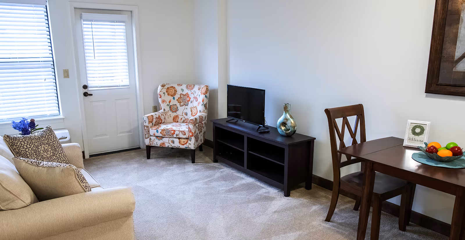 A cozy living room with a beige sofa adorned with patterned pillows, a floral armchair, a dark wooden TV stand with a flat-screen TV and decorative vase, and a wooden dining table with a chair, a bowl of fruit, and a small welcome sign. The room has light-colored walls, carpeted floor, and a door with a window covered by blinds.