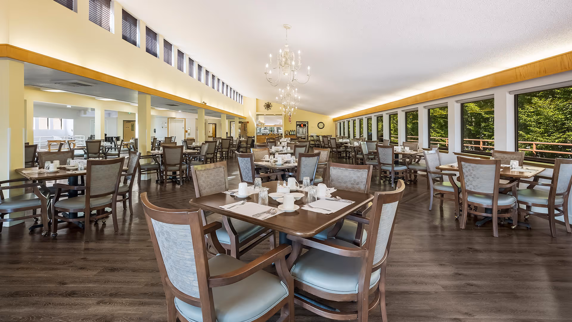 A spacious dining room with multiple wooden tables and chairs arranged neatly. Each table is set with white cups, glasses, plates, and napkins. Large windows on one side provide a view of green trees outside, and chandeliers hang from the ceiling. The room has a warm, inviting atmosphere with wooden flooring and light-colored walls.