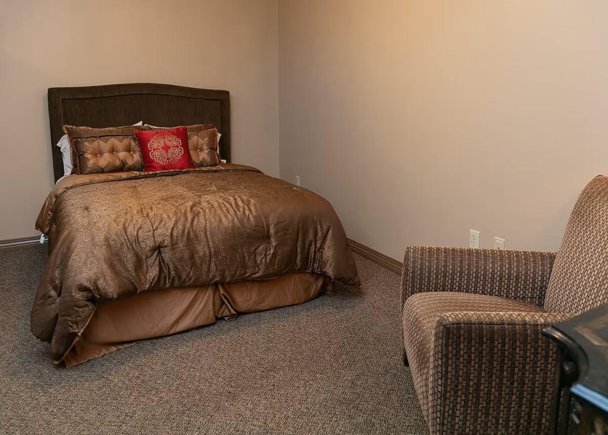 A bedroom with a brown bedspread and decorative pillows on a bed with a dark headboard. There is a patterned armchair to the right and beige walls with carpeted flooring.