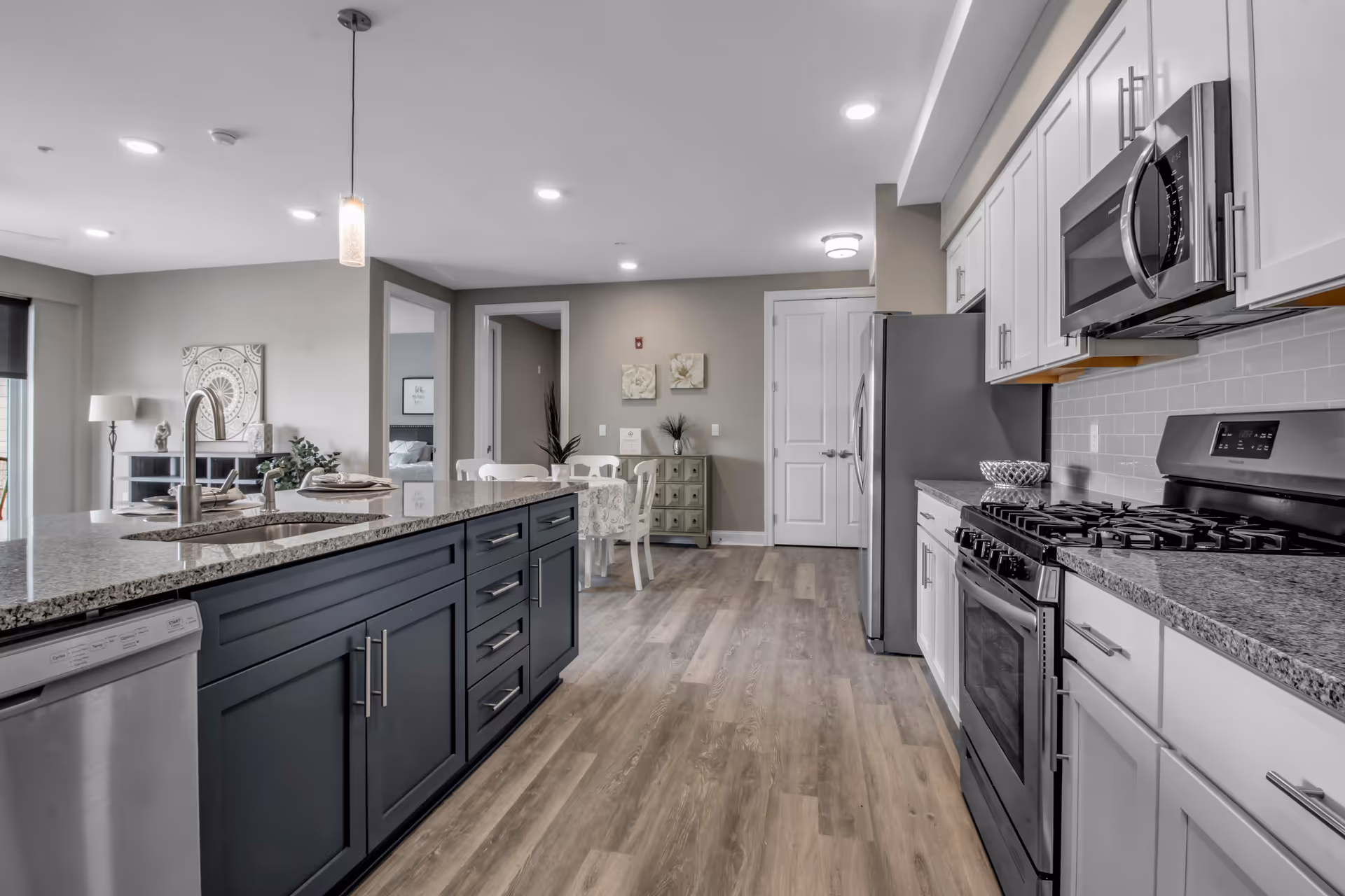 Modern kitchen with a large island featuring a granite countertop and sink, stainless steel dishwasher, stove, microwave, and refrigerator. The kitchen opens into a dining area with a table and chairs, light wood flooring, and neutral-colored walls with decorative artwork.