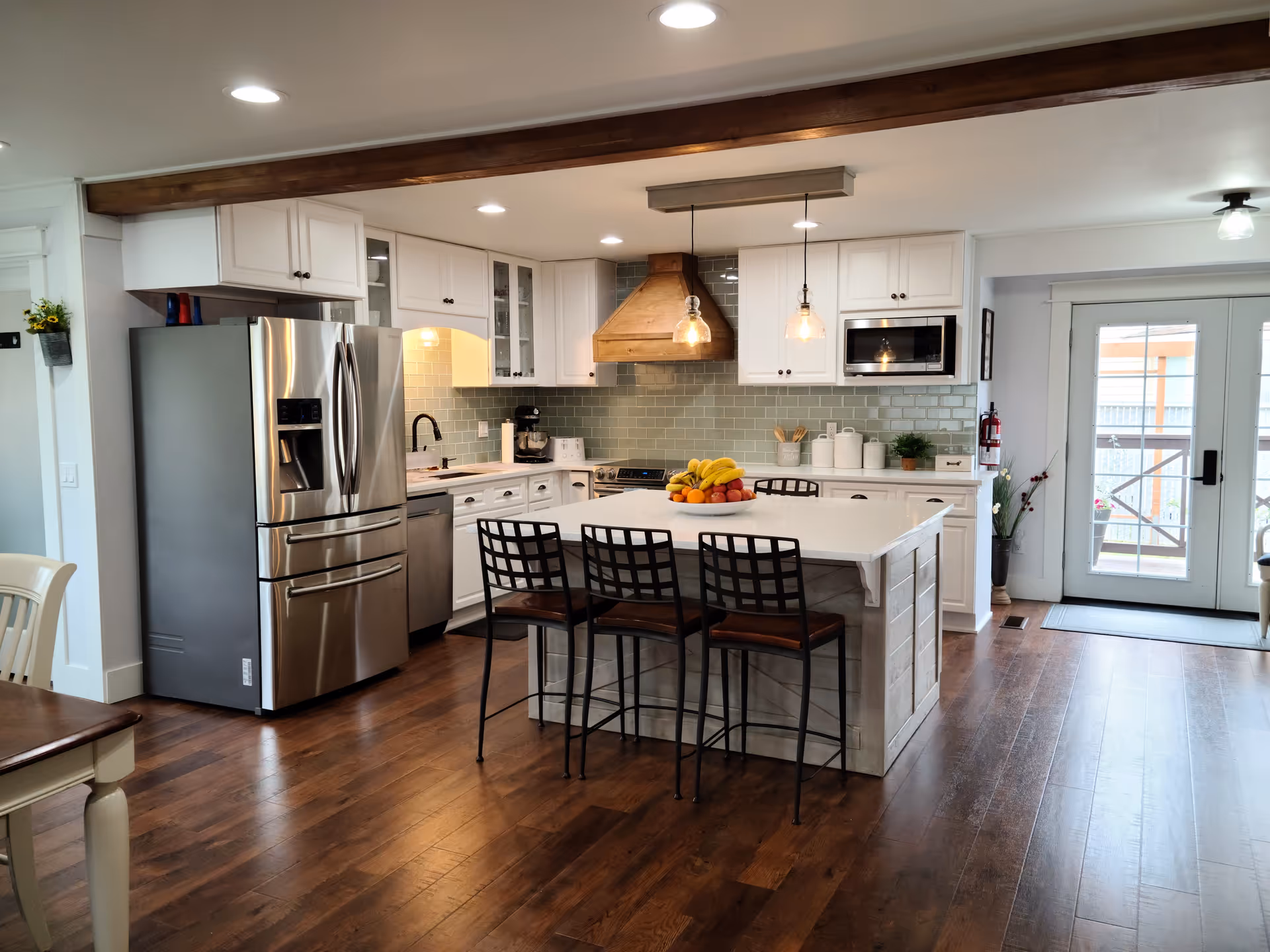 A modern kitchen with white cabinetry, a large island with three black metal chairs, stainless steel refrigerator and dishwasher, wooden floor, and pendant lights hanging above the island. There is a bowl of fruit on the island and double glass doors leading outside.