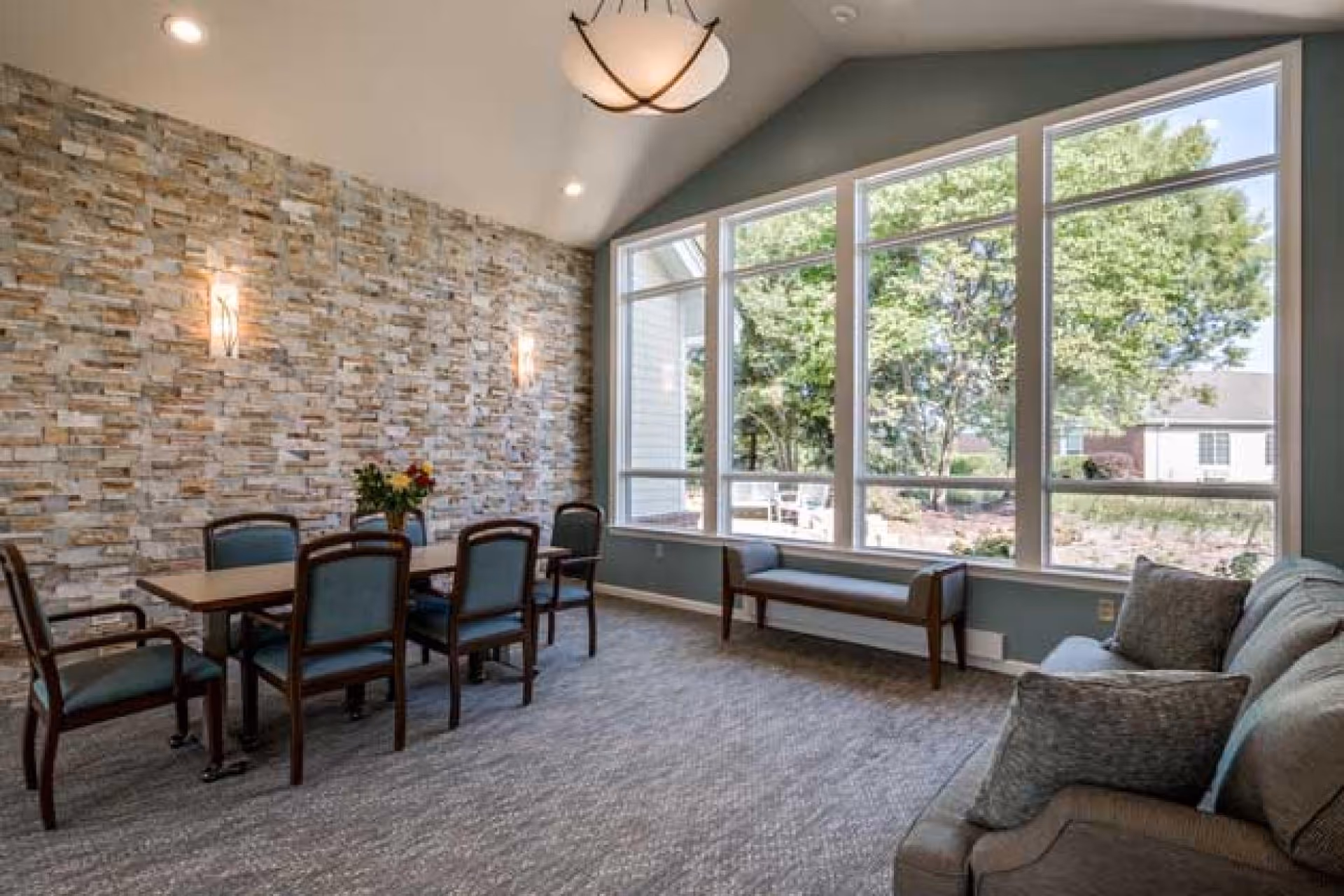A bright room with a large window showing green trees outside. Inside, there is a rectangular table with six chairs around it, a bench under the window, and a sofa with cushions. The wall behind the table has a textured stone design with two wall sconces, and a ceiling light fixture hangs above.