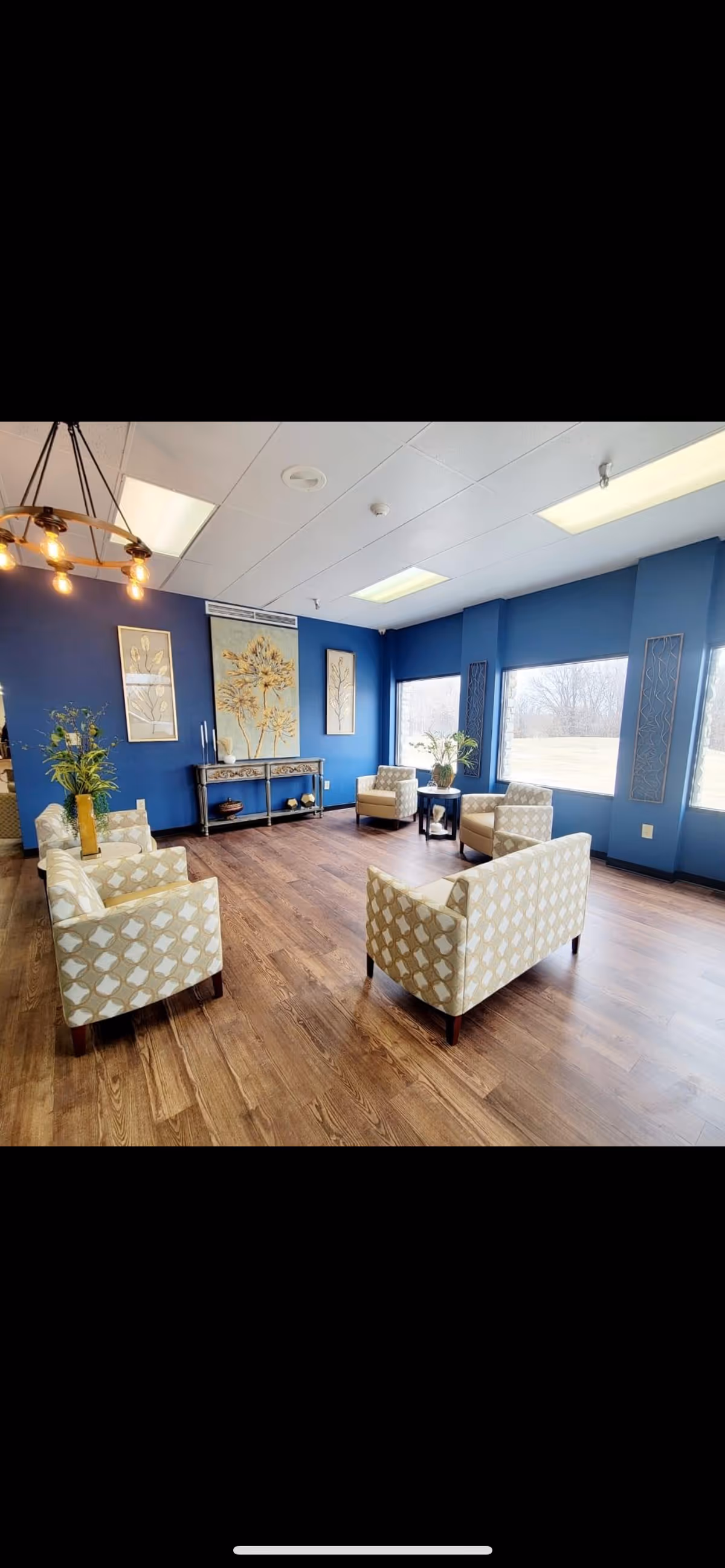 Bright communal sitting room with patterned armchairs on wood floors, blue walls, large windows and decorative wall art.