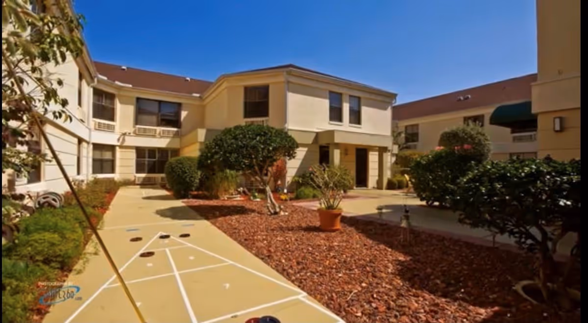 Outdoor courtyard area of a senior living facility with a shuffleboard court, surrounded by beige two-story buildings, trimmed bushes, potted plants, and a clear blue sky.