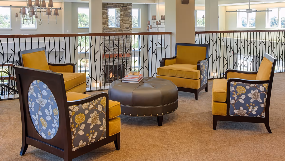 A cozy seating area with four yellow upholstered armchairs featuring floral patterned sides arranged around a round, tufted black ottoman with books on top. The area overlooks a stone fireplace below, with large windows letting in natural light and decorative black metal railings with a branch design.