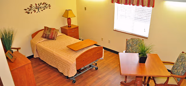 A small, neatly arranged bedroom in a skilled nursing facility featuring a single hospital-style bed with a beige quilt, a wooden bedside table with a lamp, a wooden dresser with a plant, and a wooden table with two upholstered chairs near a window with blinds and a striped valance.