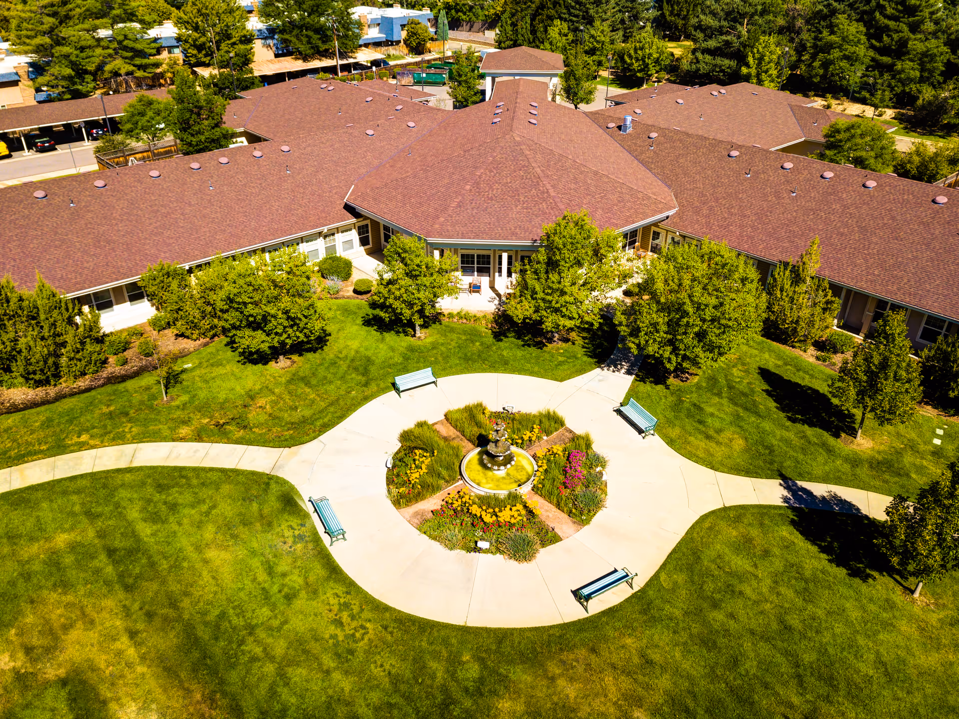 Aerial view of a senior living facility's landscaped central courtyard with a circular fountain, walkways, benches, and surrounding one-story buildings.