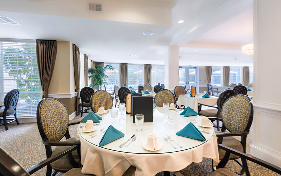 Bright dining room with round tables covered in white tablecloths, set with green folded napkins, glassware, and white cups. Upholstered chairs with patterned backs surround the tables. Large windows with brown curtains allow natural light to fill the room.