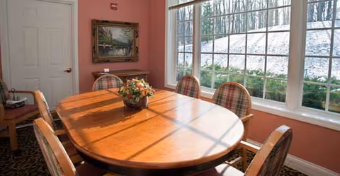 A dining room with a wooden oval table surrounded by six cushioned chairs. A floral centerpiece is placed on the table. The room has large windows letting in natural light, with a view of a snowy landscape outside. A painting hangs on the wall next to a closed white door.