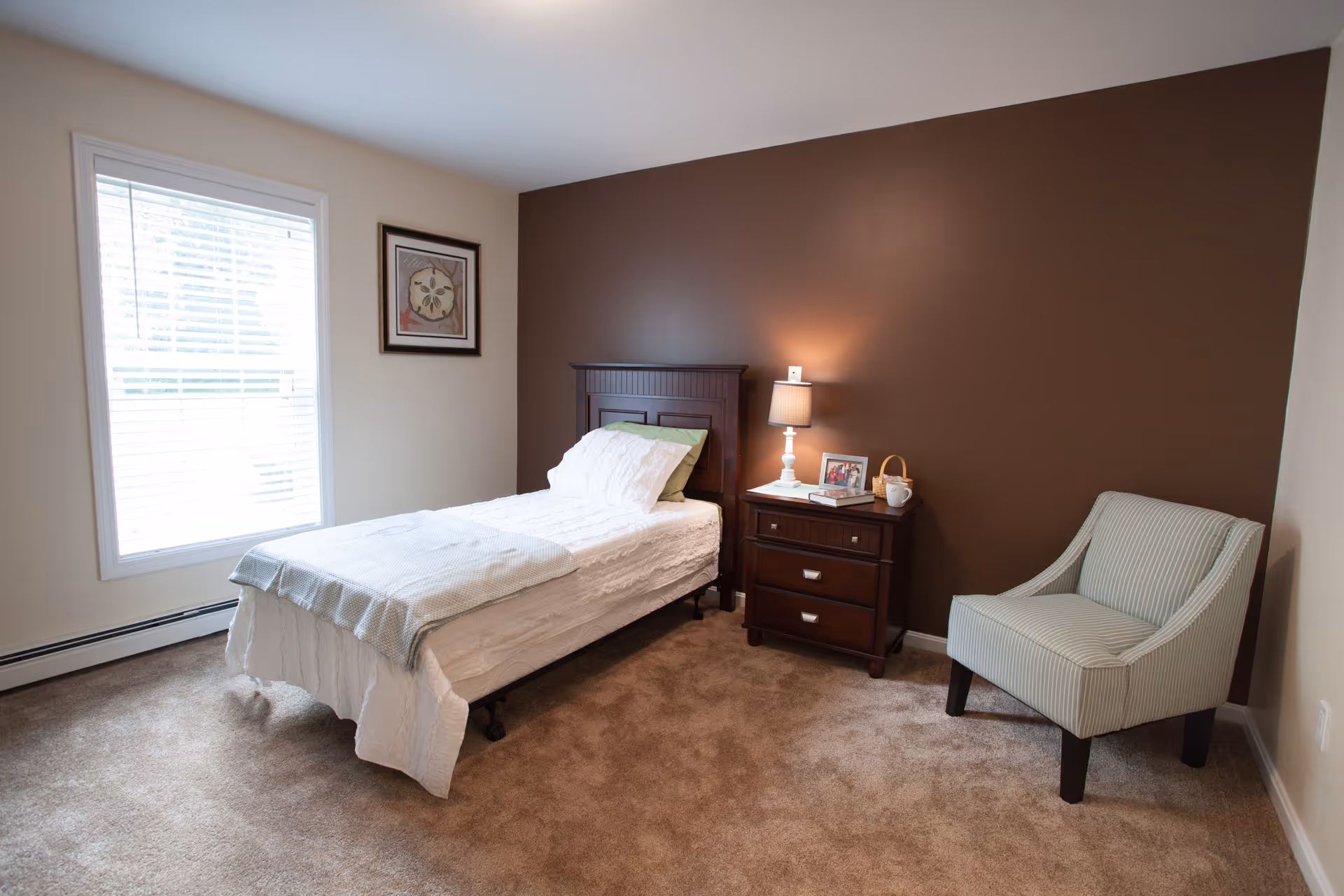 A simple bedroom with a single bed covered in white bedding, a wooden headboard, a nightstand with a lamp, framed photo, and a cup, a striped armchair, beige carpet, and a window with blinds letting in natural light.