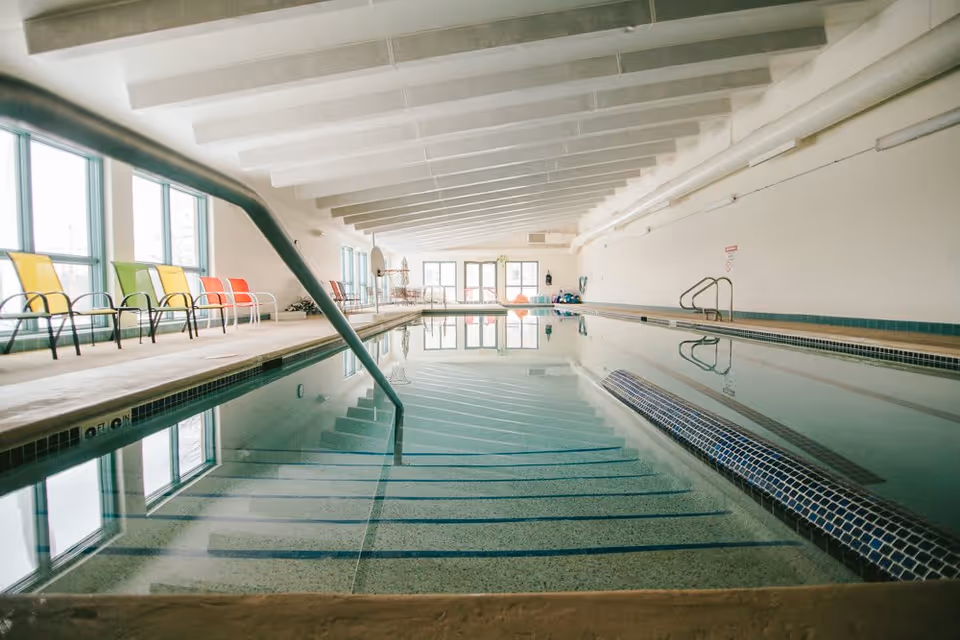 Indoor swimming pool with clear water, metal handrails, and steps leading into the pool. Colorful chairs line the left side near large windows, and the ceiling has exposed beams. The pool area is well-lit with natural light coming through the windows.