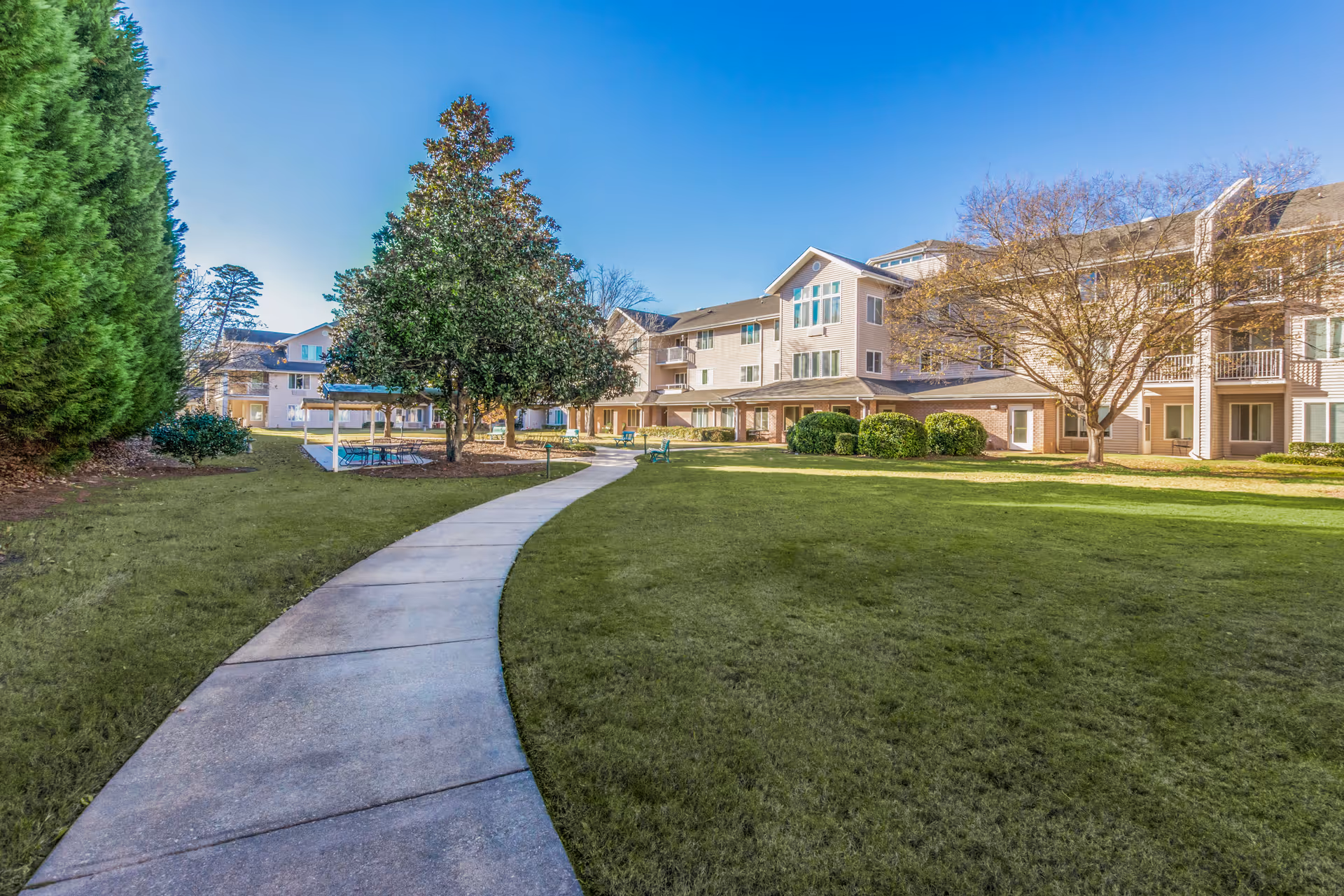 A paved walkway curves through a grassy courtyard area with trees and benches, leading to a covered seating area with tables and chairs. Surrounding the courtyard are multi-story residential buildings under a clear blue sky.