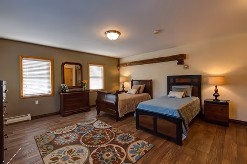 A cozy bedroom with two single beds, one with a wooden sleigh bed frame and the other with a dark wood headboard and footboard. The room features wooden flooring, a colorful patterned area rug, a wooden dresser with a mirror, two windows with blinds, and two bedside tables with lamps. The walls are painted in neutral tones with a wooden beam accent above the beds.