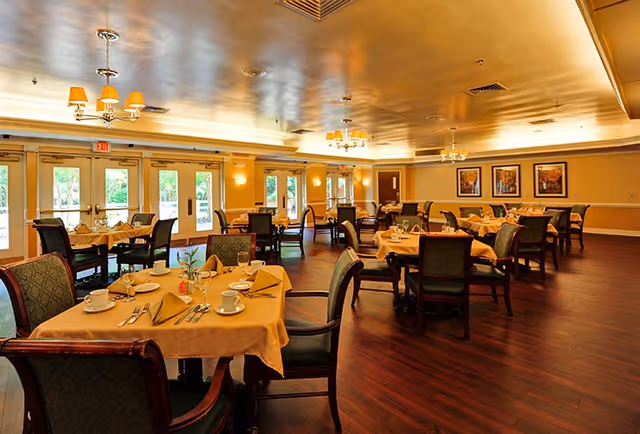 A spacious dining room with multiple tables covered in beige tablecloths, each set with cups, plates, silverware, and napkins. The room has wooden flooring, cushioned chairs, large windows and glass doors letting in natural light, and warm ceiling lighting fixtures. There are framed pictures on the far wall.