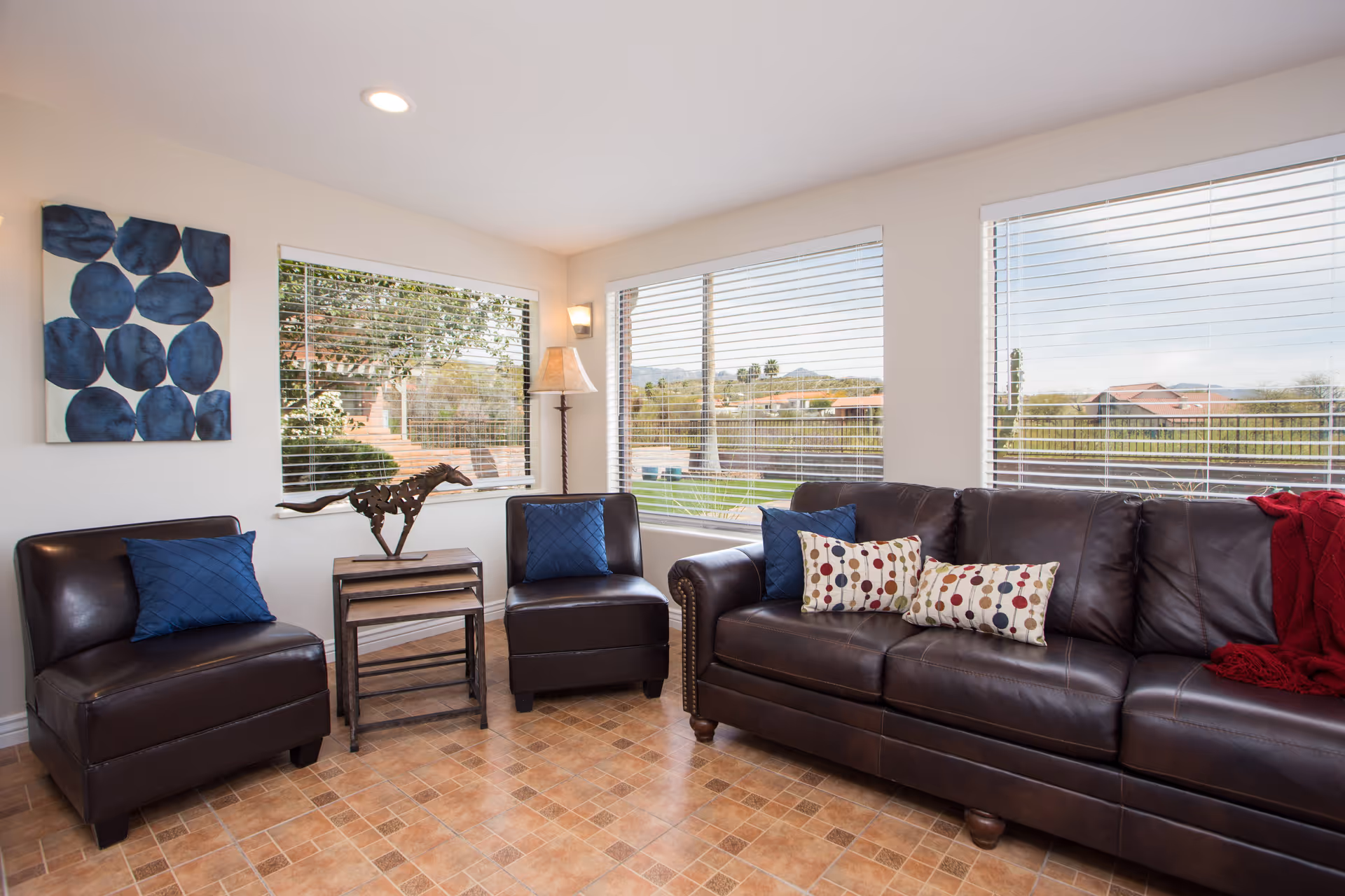A bright living room with large windows showing an outdoor view. The room features a dark brown leather sofa with colorful pillows and a red throw blanket, two matching leather chairs with blue pillows, a set of nested wooden tables with a horse sculpture on top, a floor lamp, and a blue abstract painting on the wall.