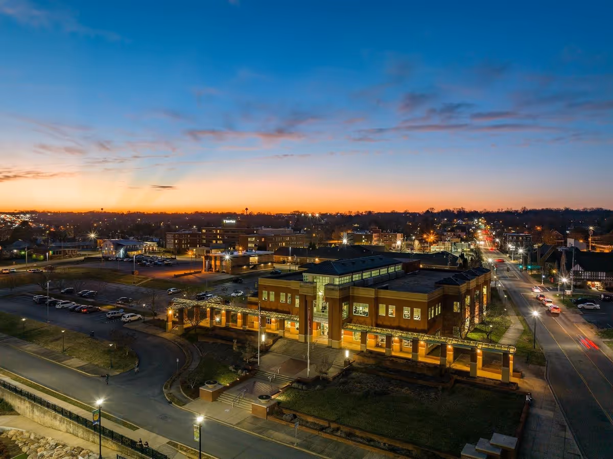 Aerial view of a town at dusk with a large brick building in the foreground illuminated by exterior lights. The sky is clear with a gradient from orange near the horizon to blue higher up. Streets with cars and streetlights are visible, along with other buildings and parking lots in the background.