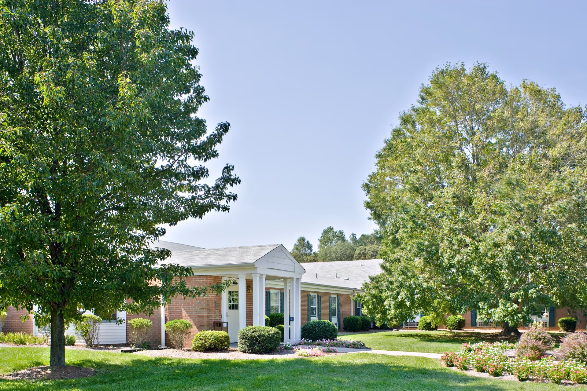 Exterior view of a single-story brick building with white columns at the entrance, surrounded by green trees, bushes, and a well-maintained lawn under a clear blue sky.
