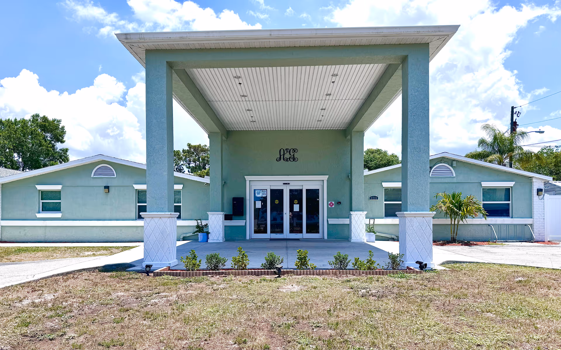 Front exterior view of a single-story building with a large covered entrance supported by two columns. The building is painted light green with white trim and has several windows. There are small bushes planted in front of the entrance and a clear blue sky with some clouds in the background.