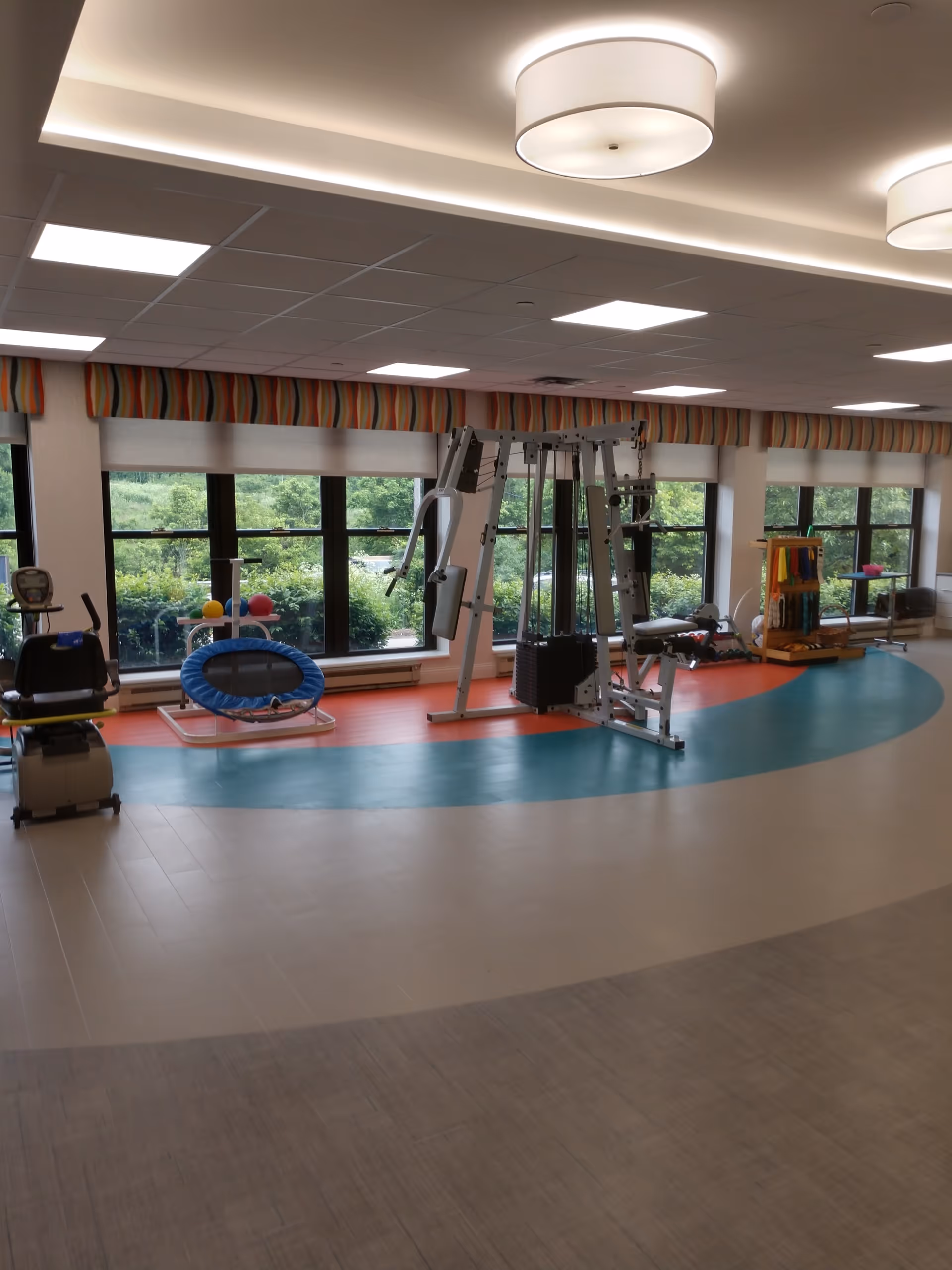 A bright and spacious rehabilitation gym area with exercise equipment including a multi-station weight machine, a recumbent bike, a small trampoline, and various therapy tools. Large windows with colorful valances let in natural light and show greenery outside.