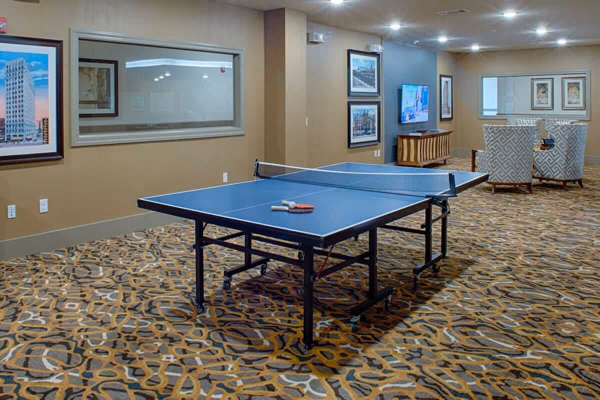 A spacious indoor recreational room with a blue ping pong table in the center, two paddles and a ball on the table, patterned carpet flooring, beige walls adorned with framed pictures, a flat-screen TV mounted on the wall, and two patterned armchairs with a small table in the background.