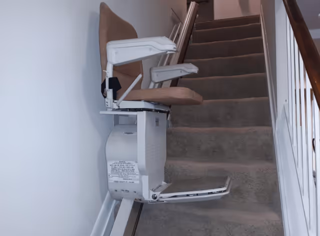 A beige stairlift chair installed on a rail along a carpeted indoor staircase.