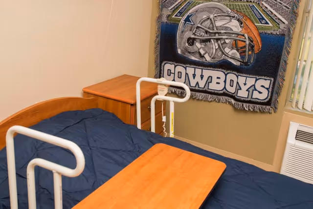 A small bedroom with a hospital-style bed covered with a dark blue blanket, a wooden bedside table, and a wooden overbed table. A Dallas Cowboys blanket with a football helmet and stadium design hangs on the wall above the bed. There is a window with vertical blinds and a wall-mounted air conditioning unit.