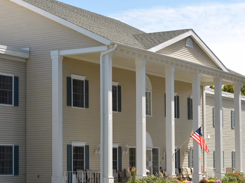 Exterior view of a beige two-story building with white columns and green window shutters. There is a porch with chairs and an American flag displayed near the entrance under a clear sky.