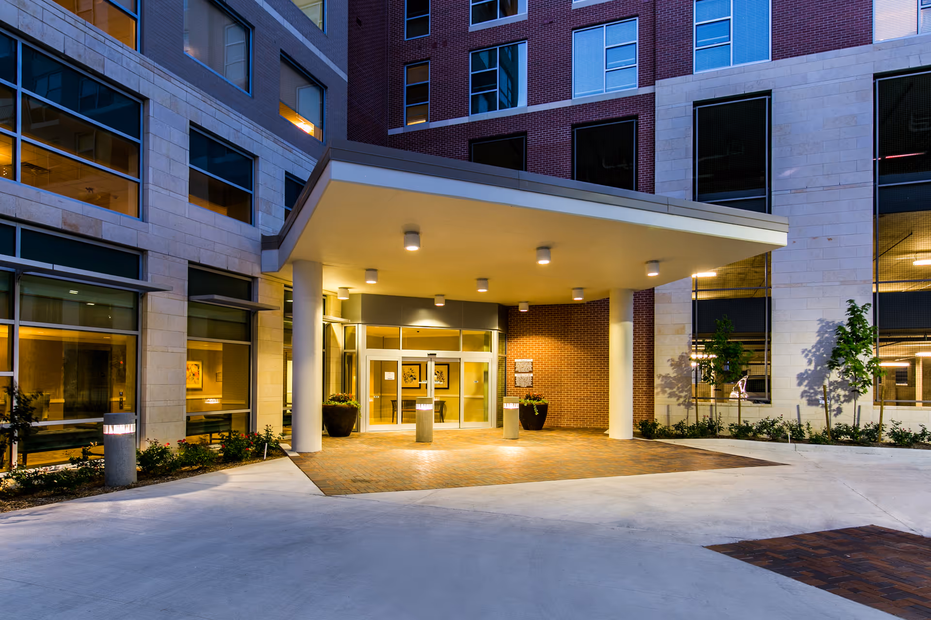 Entrance of a modern senior living facility building at dusk with a covered drop-off area, large glass doors, and illuminated exterior lights. The building features a combination of brick and stone facade with multiple windows and some landscaping around the entrance.
