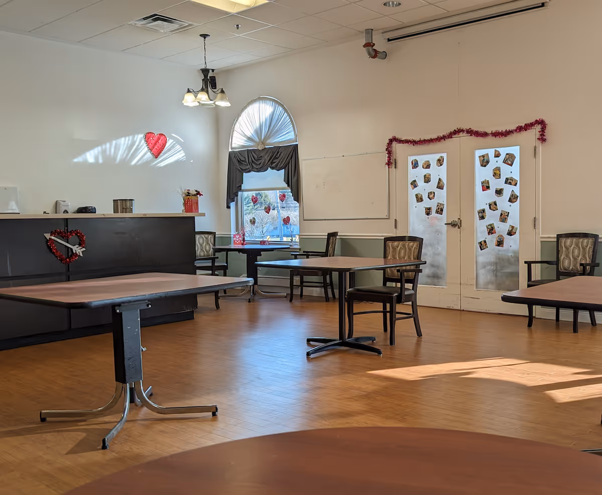 A bright room with wooden floors and several tables and chairs arranged around the space. There is a counter on the left side decorated with a heart-shaped wreath. A window with a half-circle top is adorned with curtains and heart decorations. Double doors at the back have photos and a garland hanging above them. The room is lit by natural light and a ceiling light fixture.