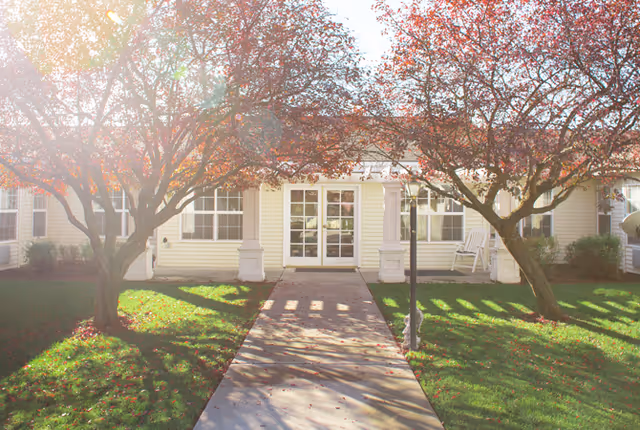 Path leading to the front entrance of a single-story senior living building framed by two trees and a manicured lawn.
