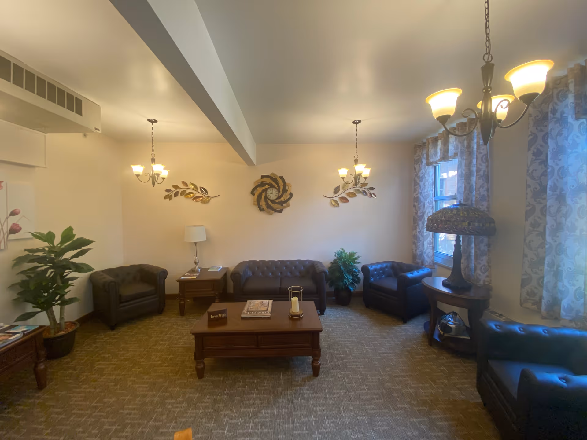 A cozy living room with brown leather sofas and armchairs arranged around a wooden coffee table. The room has beige walls decorated with metal leaf wall art and a circular wall sculpture. There are two windows with patterned curtains letting in natural light. The room is lit by three ceiling chandeliers with warm lights. There are also two potted plants and a table lamp with a stained glass shade on a side table.