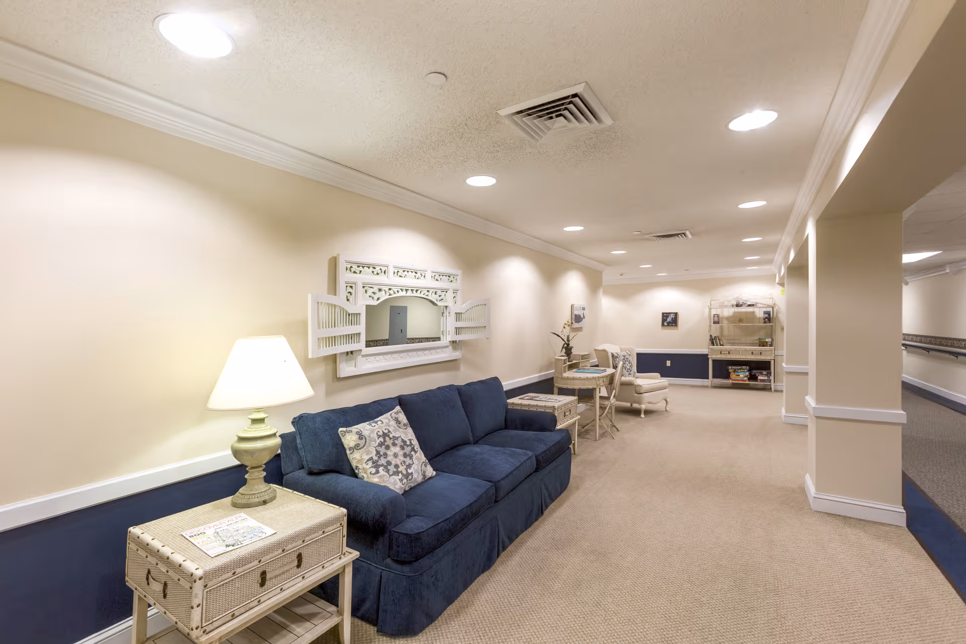 A well-lit hallway in a senior living facility featuring a blue couch with patterned pillows, a side table with a lamp, a decorative wall mirror, and additional seating with a small table further down the hallway. The walls are painted beige with a dark blue lower section, and the carpet is light beige. The hallway extends into the distance with ceiling lights and ventilation vents visible.