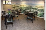 A small dining area with several black metal tables and chairs arranged on a carpeted floor. Each table has a small vase with flowers. The back wall features a large mural depicting a scenic landscape with rolling hills and greenery.