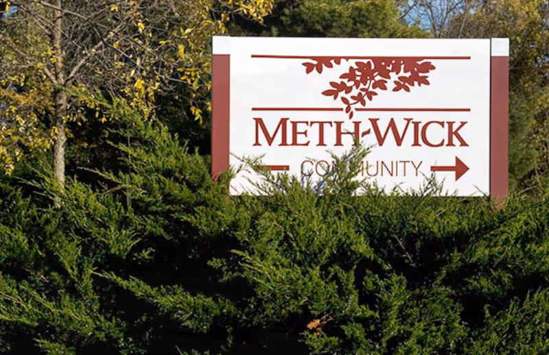 A white and maroon Meth-Wick Community sign partially obscured by shrubs and trees.