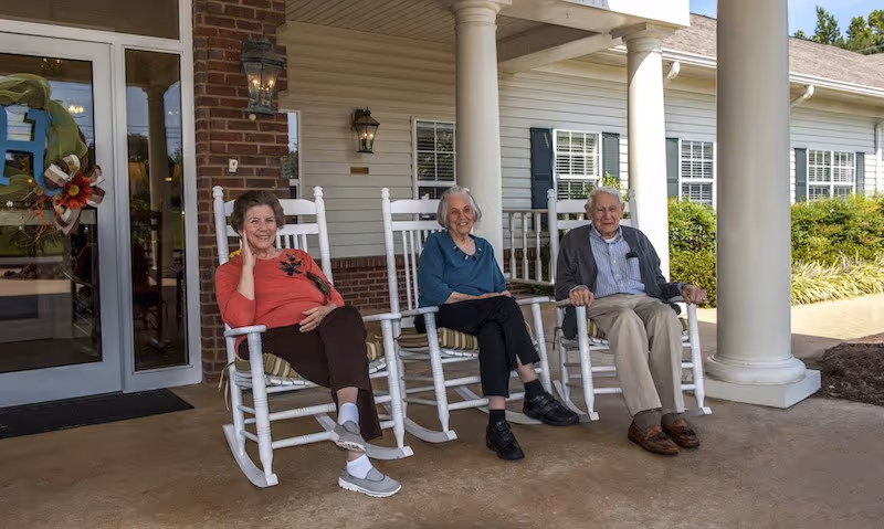 Three elderly people sitting on white rocking chairs on the porch of a building with white siding and brick accents. The porch has large white columns and a glass door with a decorative wreath. The people appear relaxed and are smiling.