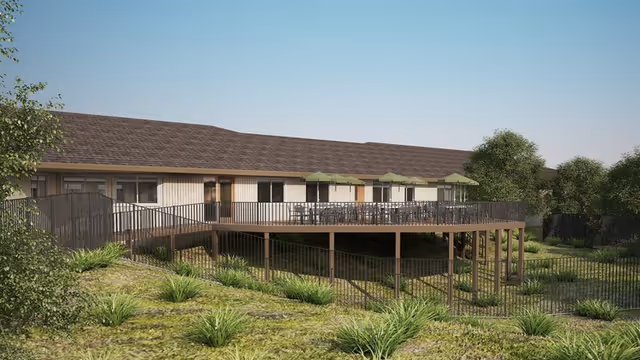 Exterior view of a single-story senior living building with a raised wooden deck, outdoor seating with umbrellas, and landscaped grounds.