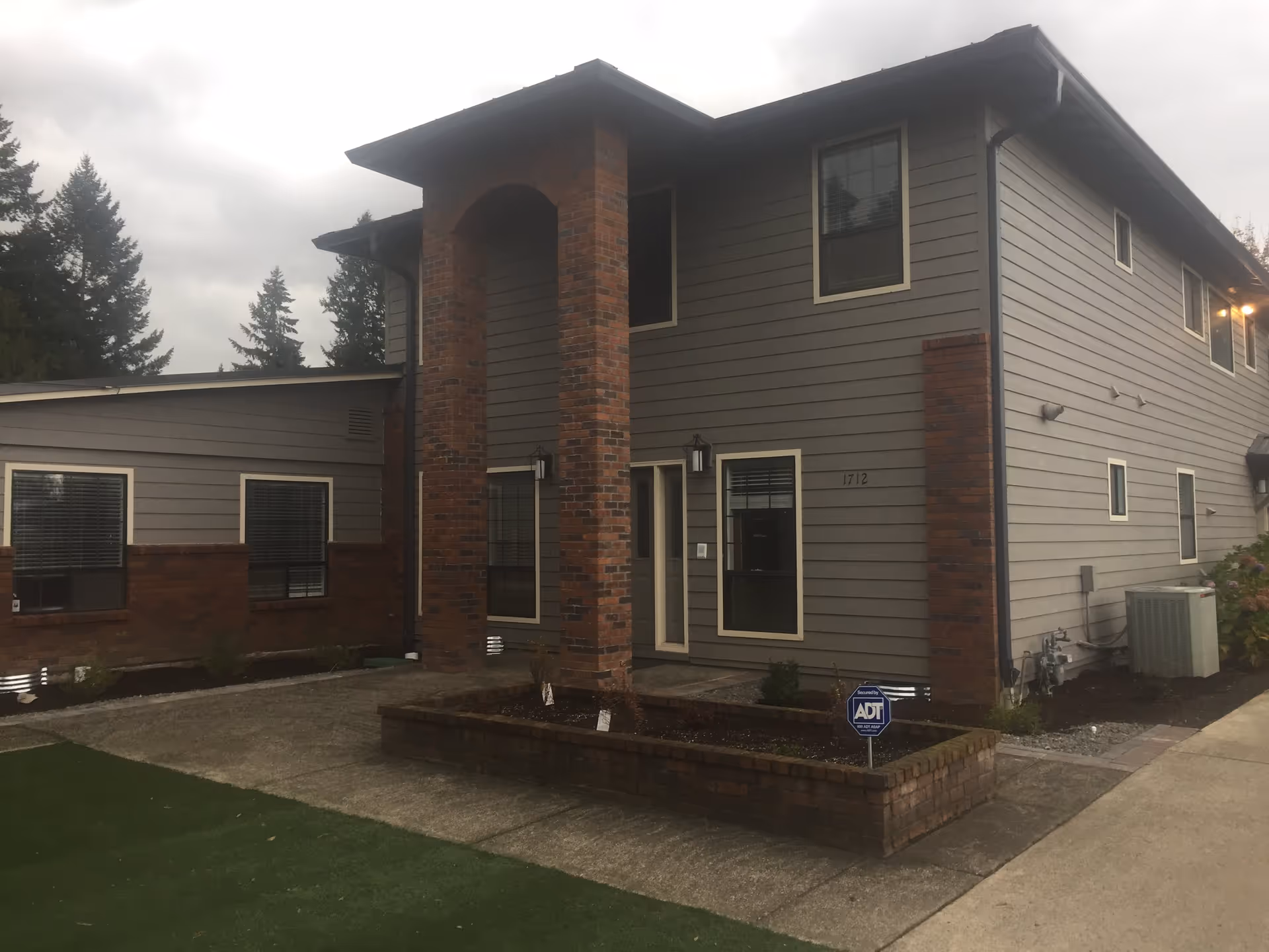 Exterior view of a two-story building with gray siding and brick accents, featuring multiple windows and a covered entrance supported by brick columns. There is a small garden bed bordered by bricks and an ADT security sign in front. The sky is overcast and there are tall evergreen trees in the background.