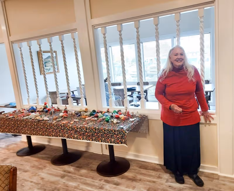 An elderly woman with long white hair wearing a red sweater and dark skirt stands smiling next to a table covered with a floral tablecloth displaying various colorful holiday ornaments. The setting appears to be an indoor community room with wooden flooring and white railings separating the space from another room with large windows.
